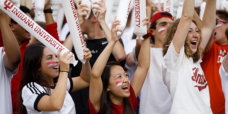 A group of students dressed in white and crimson cheer and hold up inflatable sticks that say "Indiana University."