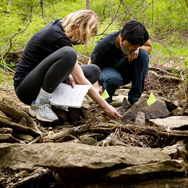 Two students holding notebooks squat to examine rocks and water at a stream outside.