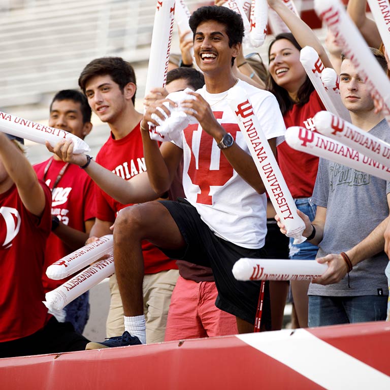 A group of students dressed in white and crimson cheer and hold up inflatable sticks that say "Indiana University." One male student has his leg up on a concrete wall.