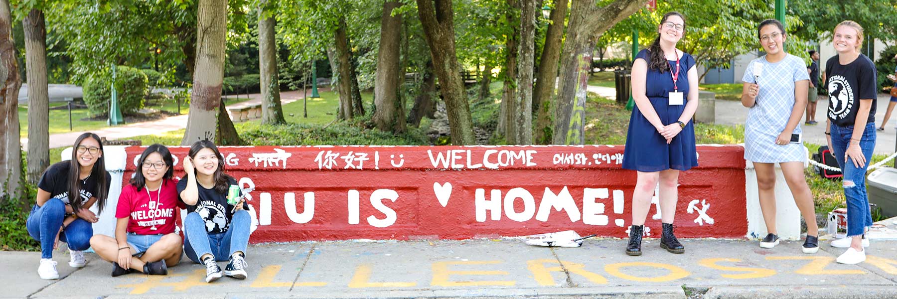 Several students in International Welcome Week tee shirts stand with paint and paintbrushes in front of a bridge wall decorated with white and red words and pictures.