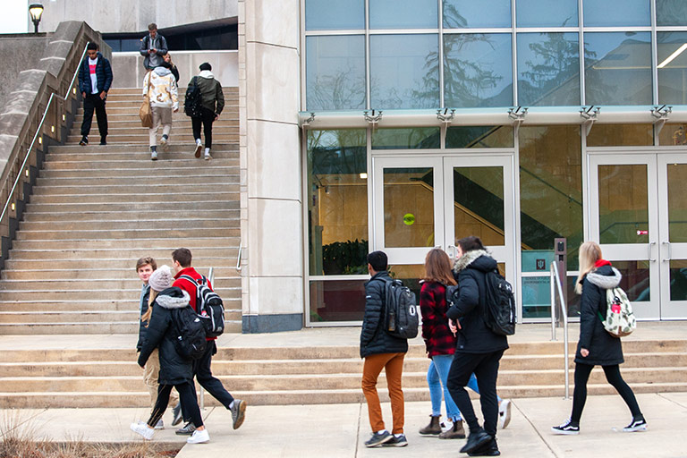 Students in winter coats walk in front and up the stairs of a campus building.