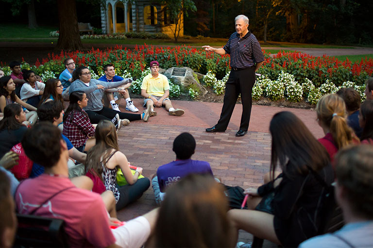 A male speaker talks to a group of students seated outside on the ground in a circle for orientation.