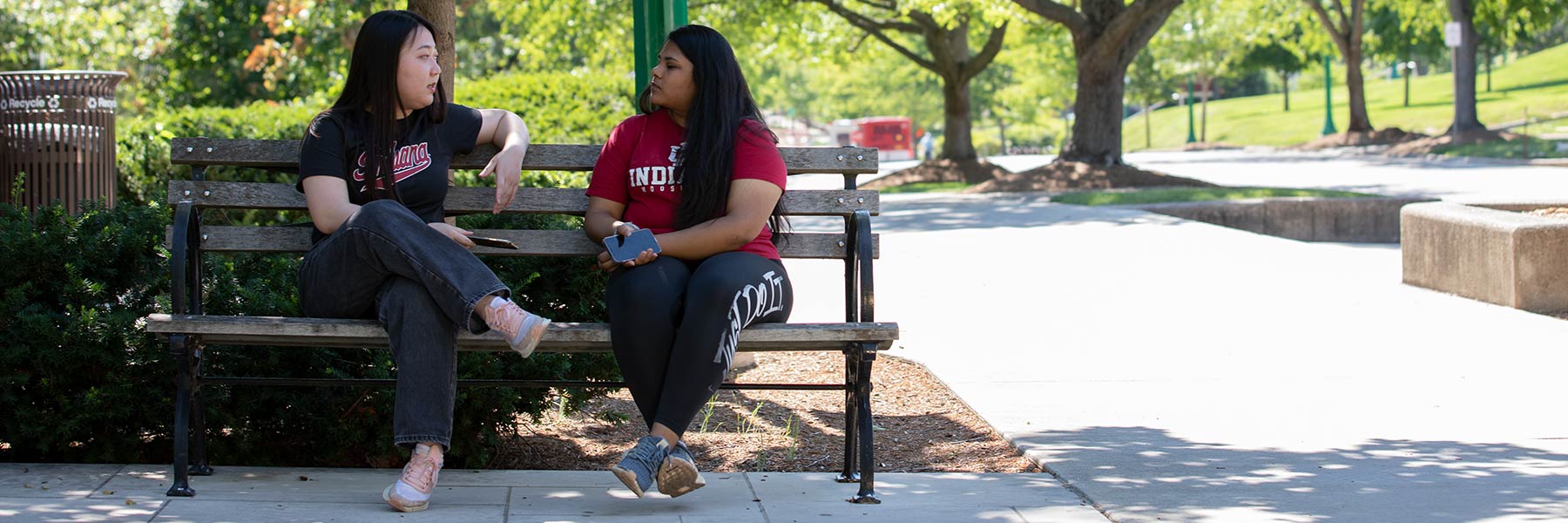 Two female students sit on outdoor benches on a sunny day talking.
