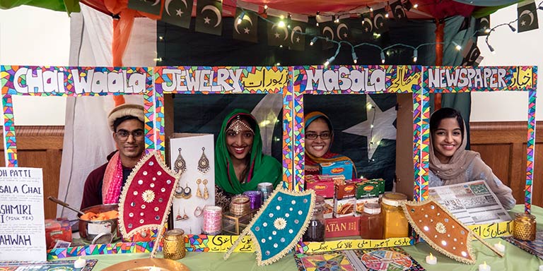 Four people sit behind homemade decorated frames that say “Chai Waala,” “Jewelry,” “Masala,” and “Newspaper” with objects sitting in front of them on a table.