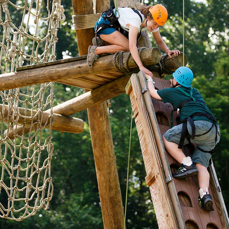 One person on a ropes course platform reaches down to help another person coming up a ladder.