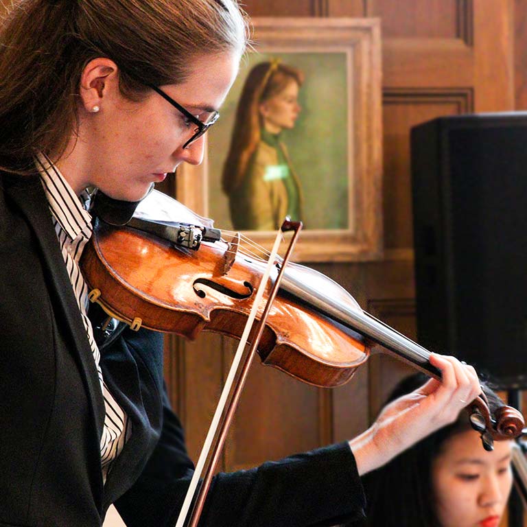A woman plays a violin; a painted portrait is visible on the wall behind her.