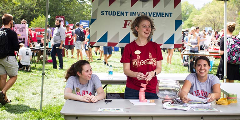 Three students stand or sit behind a welcome table with a banner that says "Student involvement fair" behind it.