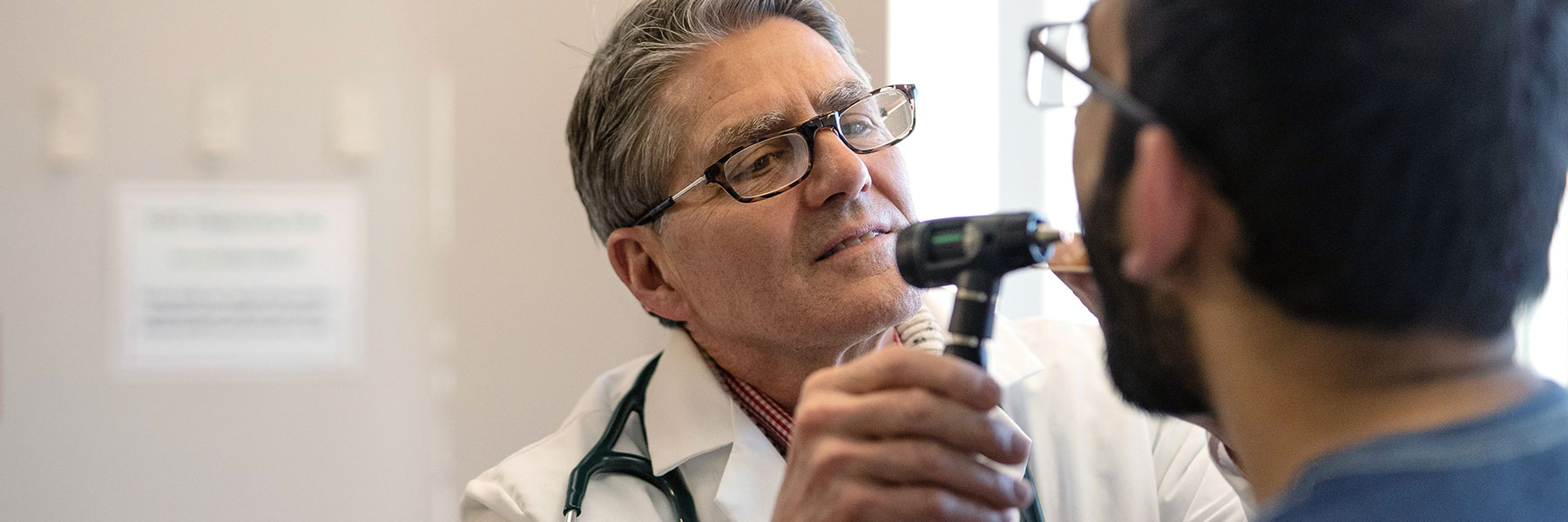 A male doctor holds up otoscope to look in the throat of a patient in front of him.