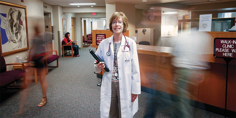 A female doctor stands in front of the health center check-in desk as blurred figures of patients walk past her.