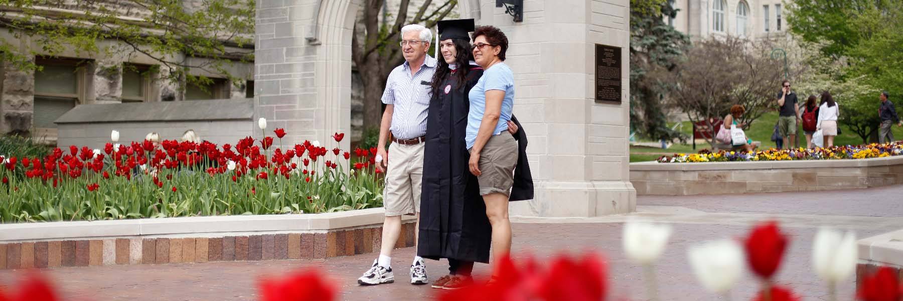 Parents stand on either side of a graduate and pose for a photo at the Sample Gates.