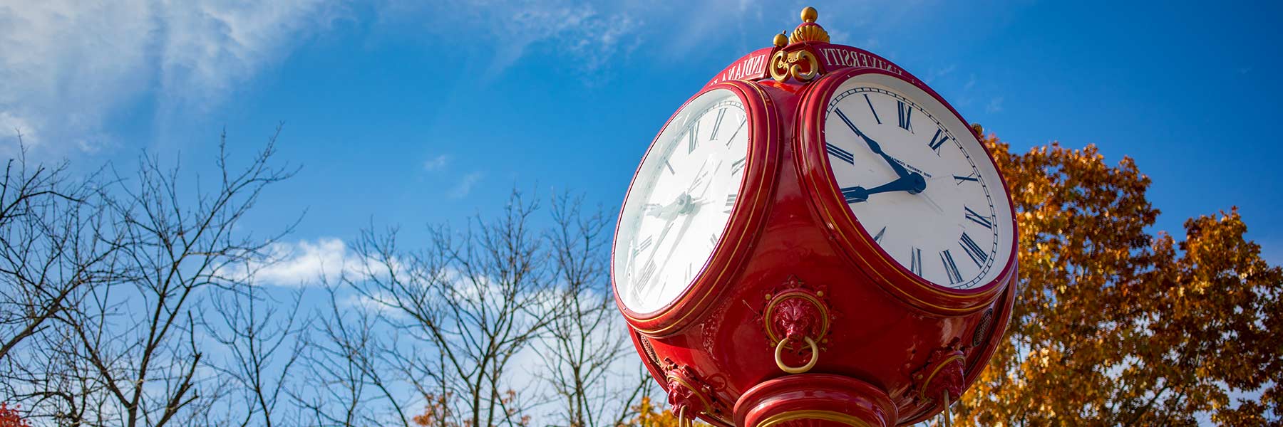 Looking up at ornate red metal clock with fall leaves and sky in the background.
