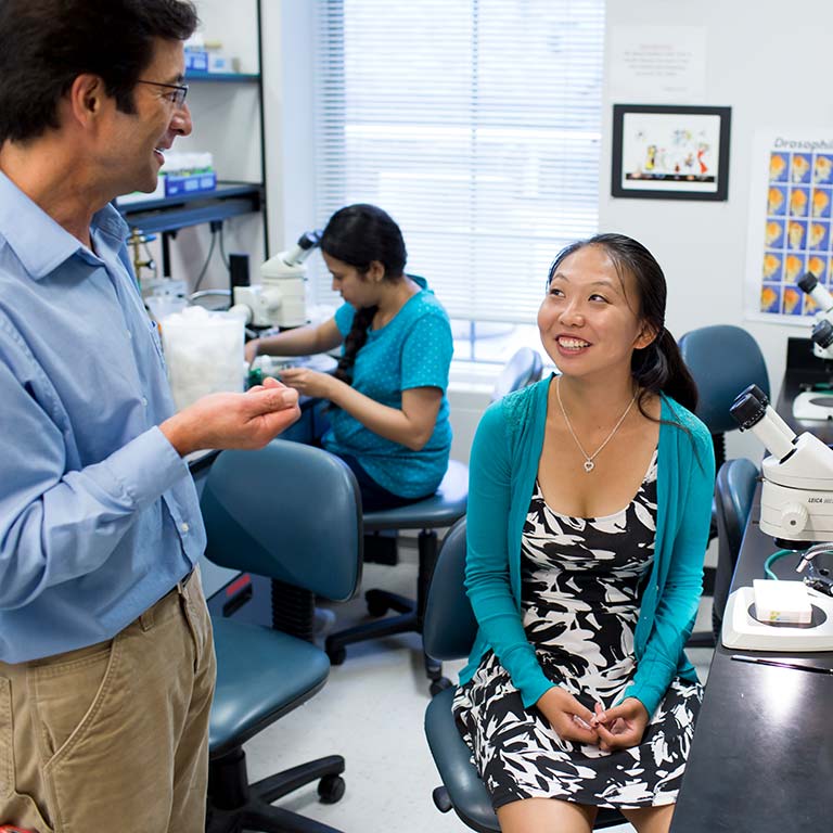 A male speaks with a woman sitting at a lab seat in front of a microscope.