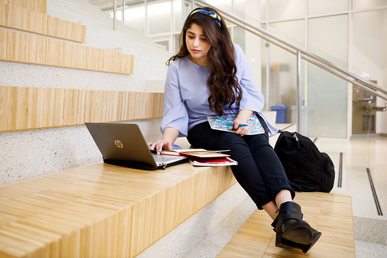 A female student sits on a wooden stairstep bench with a laptop and books in front of her.