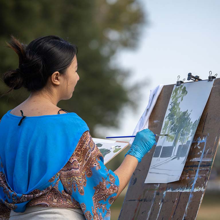 Looking over the shoulder of a female student as she paints a tree and building on an art easel.