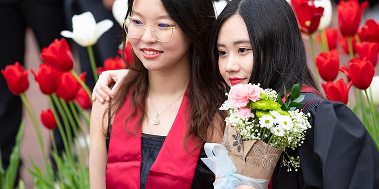 Two female graduates in commencement stoles while posing for a photo; one holds a bouquet of flowers.